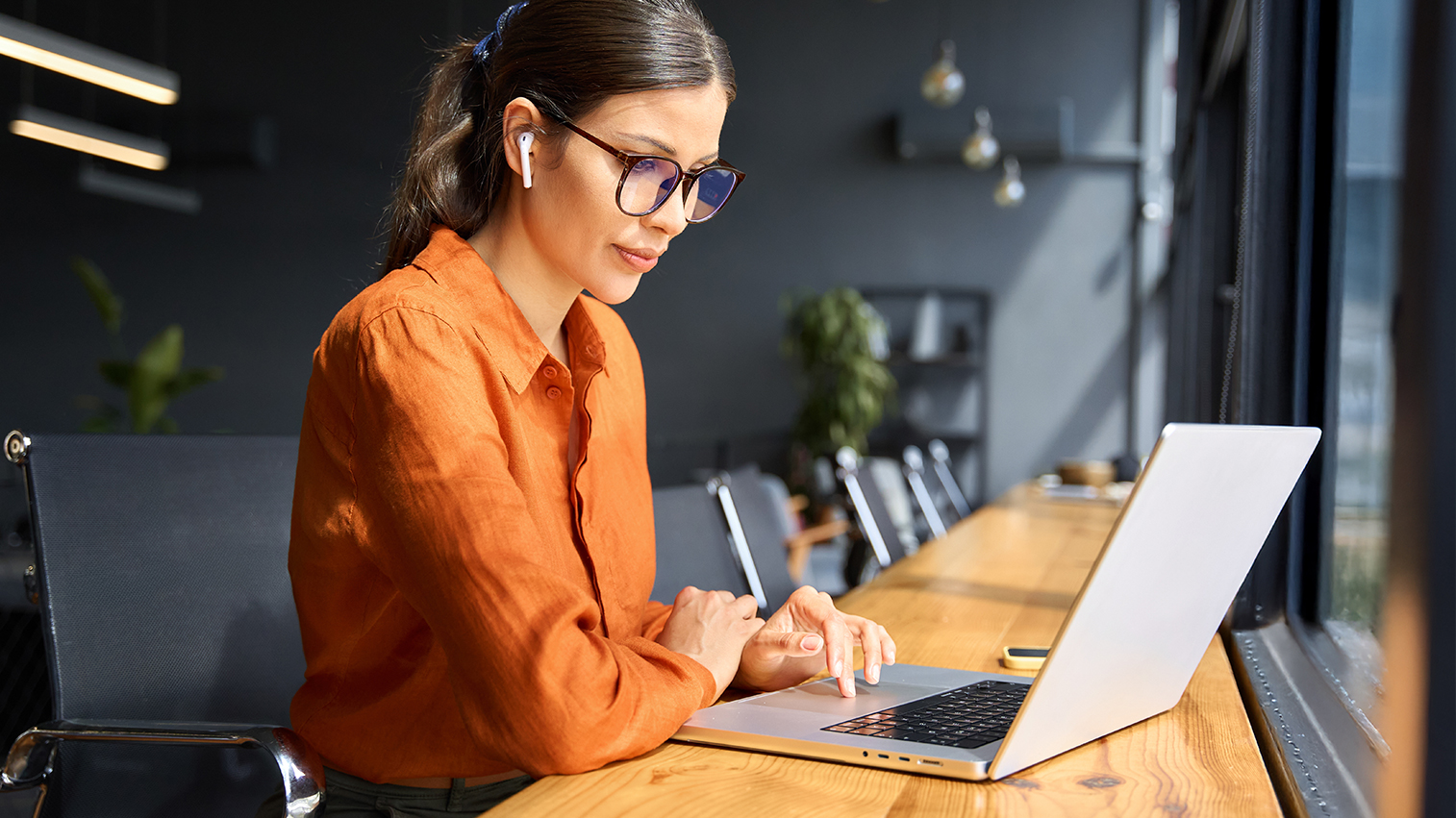 A young woman in glasses, wearing a professional orange blouse sits at a desk working on her laptop.