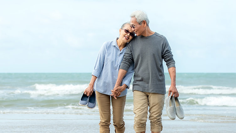 Older couple holding hands and shoes while walking along the beach